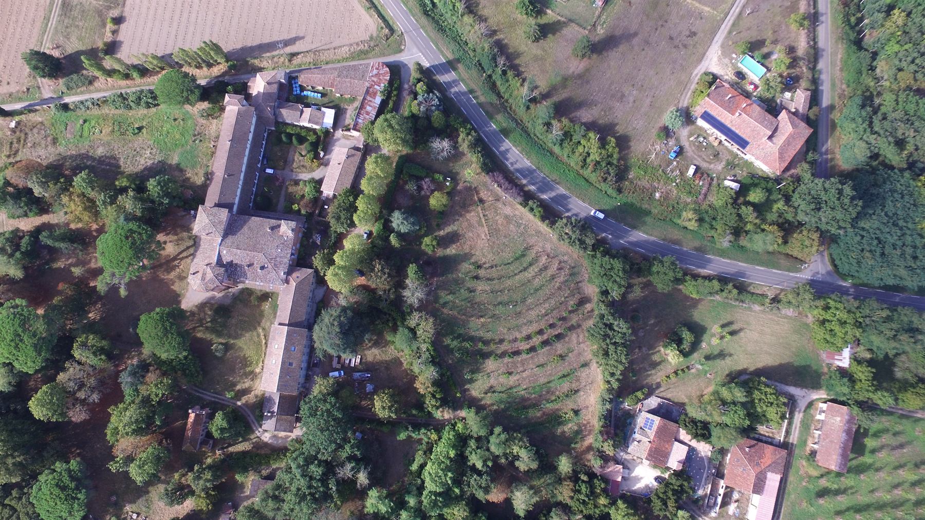 The estate from the air: abbey buildings, orchard, tree belts and open fields, with the village of Mas-Grenier beyond.