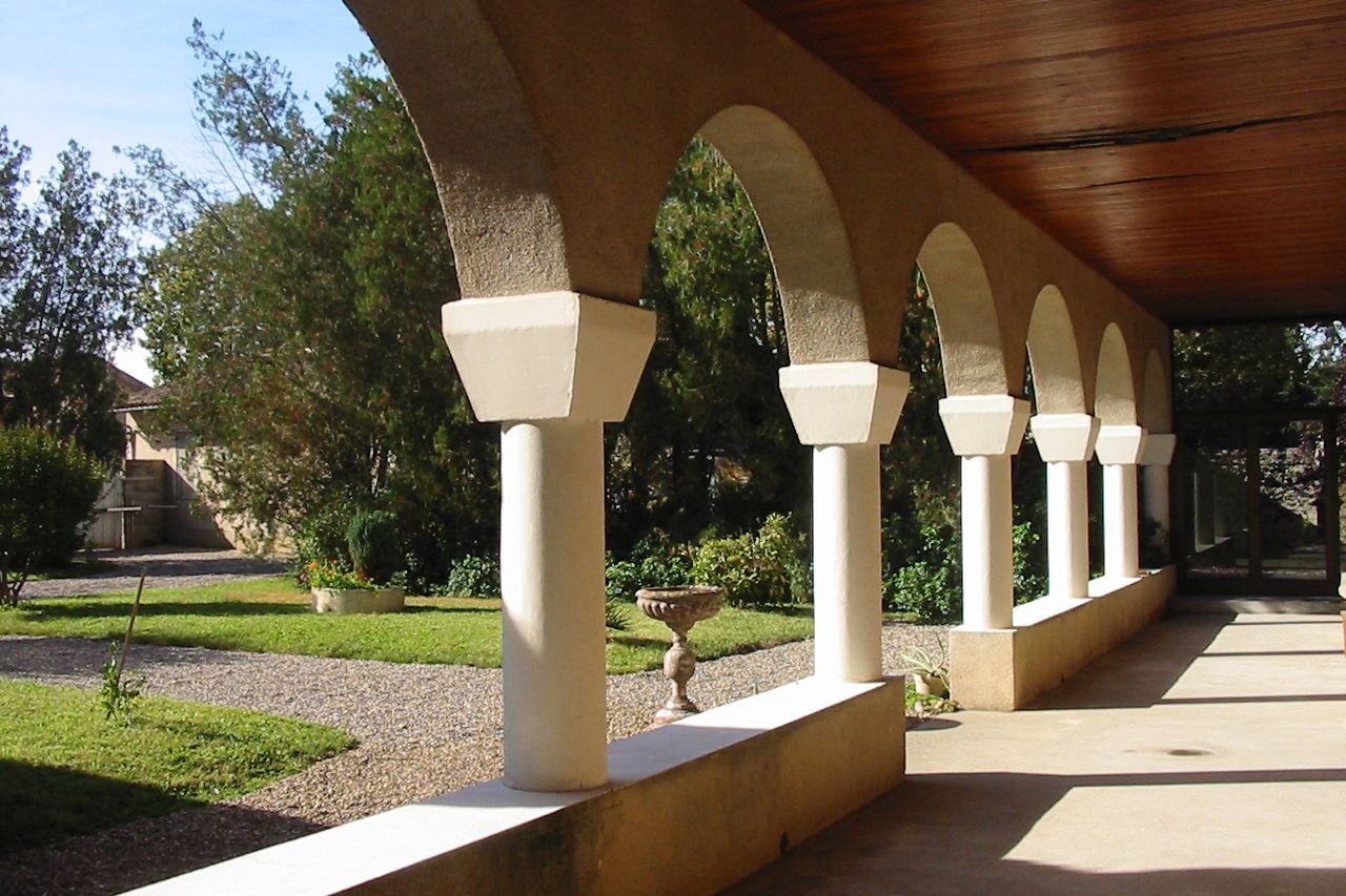 The cloister gallery: a run of round arches on square columns, opening onto a sunlit garden with a stone birdbath.