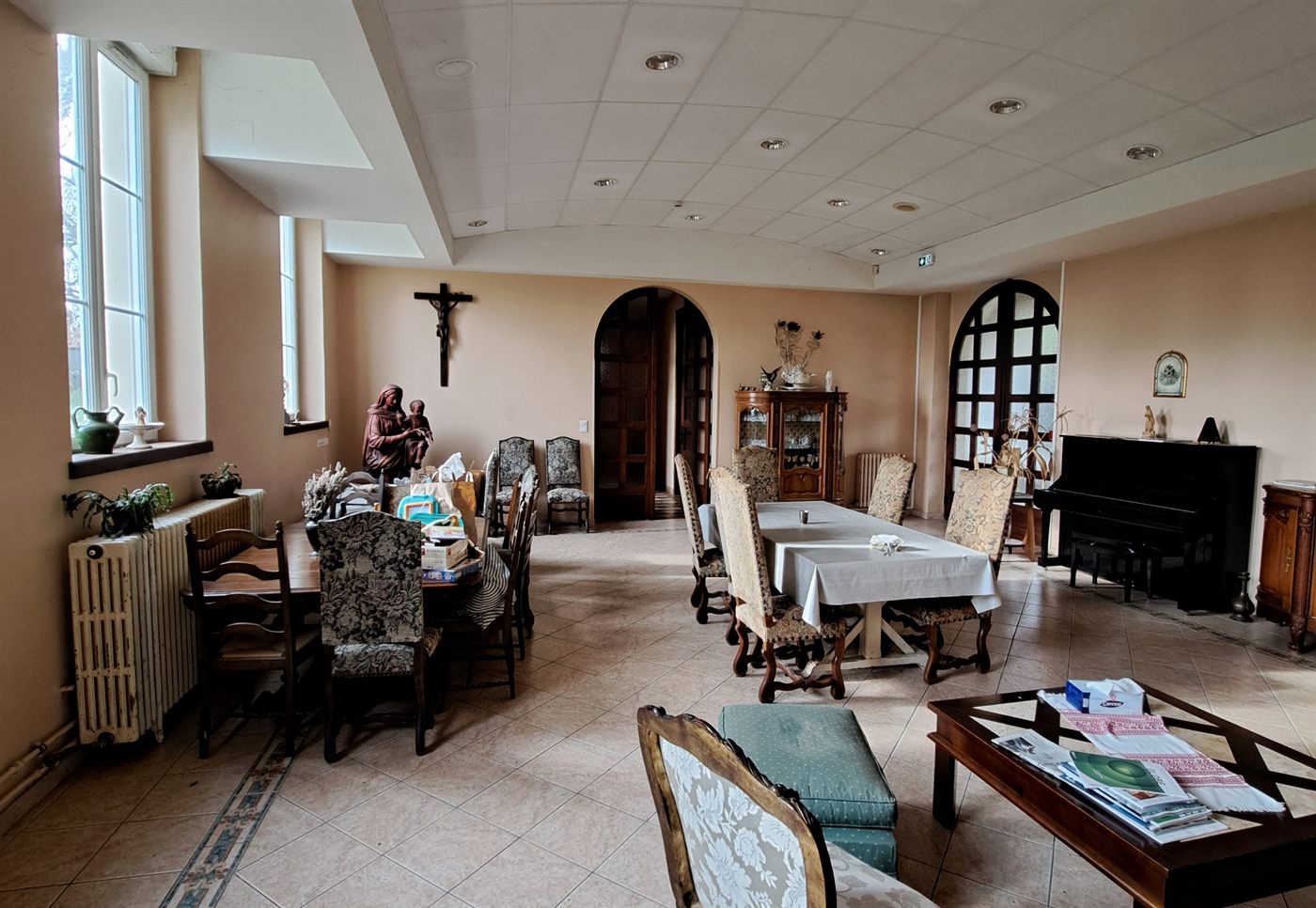 The refectory: long tables laid, tapestry chairs, an upright piano and an arched doorway at the far end.