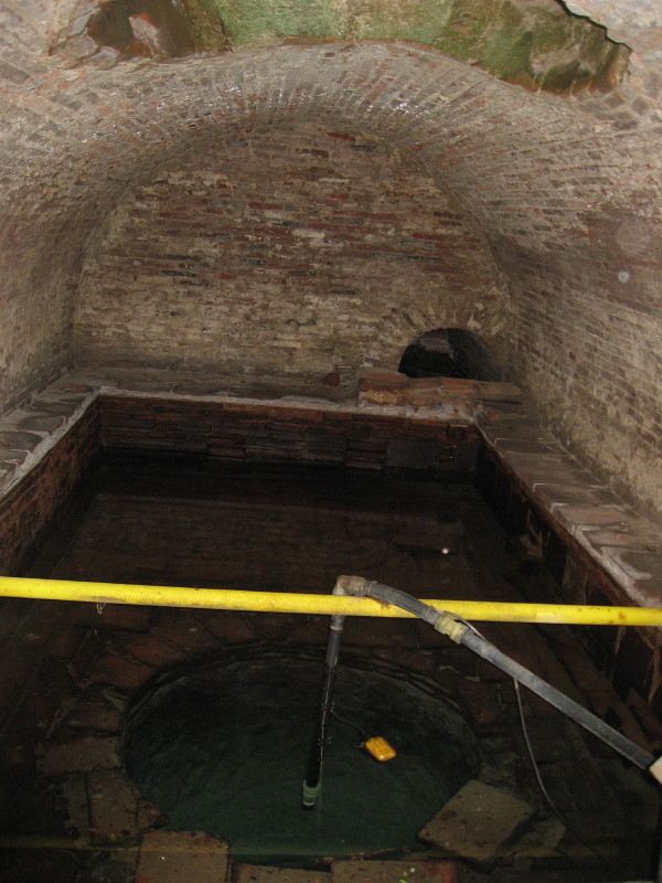 The vaulted brick cistern of the abbey spring: an old brick barrel vault over a basin of dark standing water, crossed by a modern intake pipe.